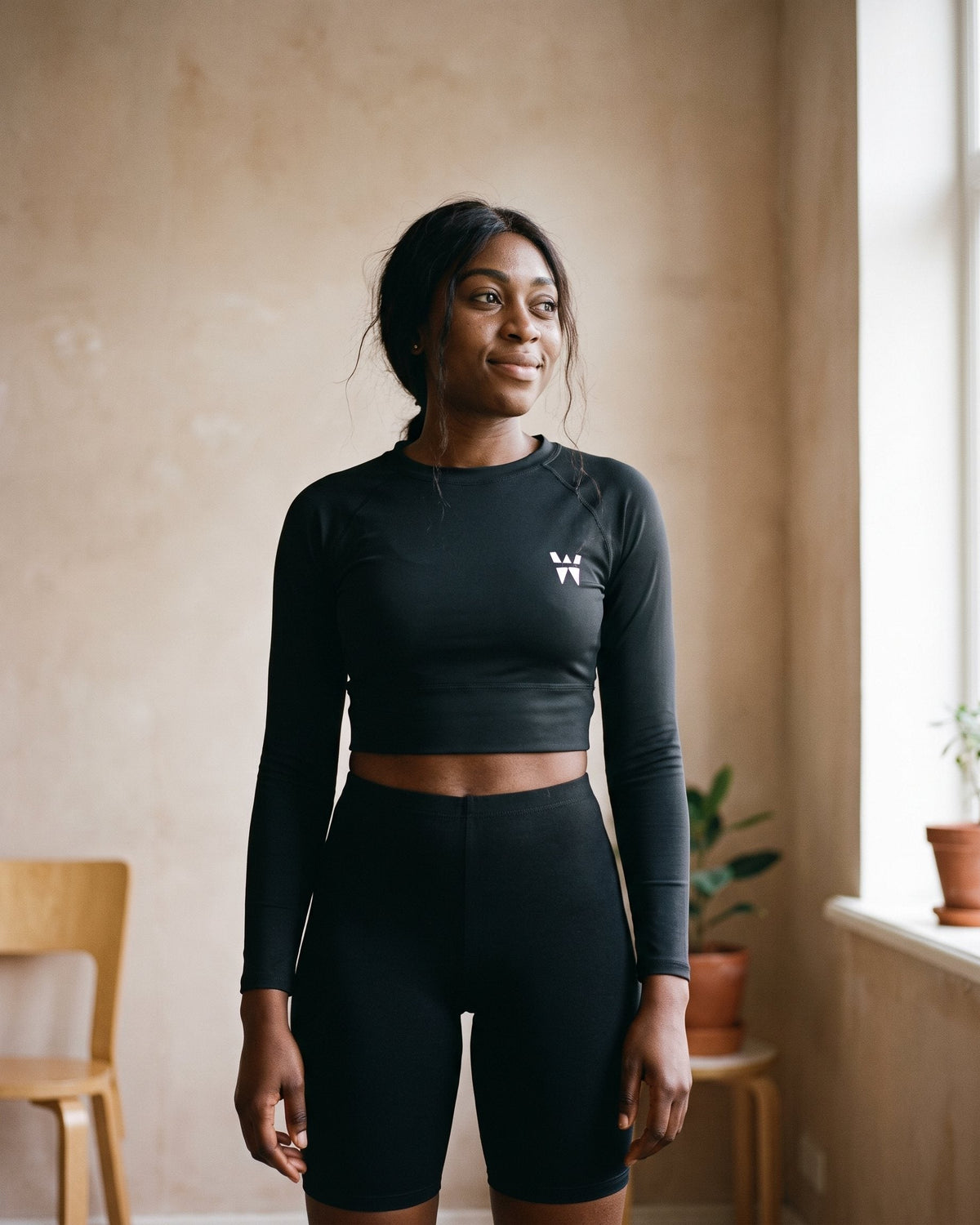 A woman in a plaster-walled room wearing the WAYSHNER Vapour Long-Sleeve Crop and Sculpt Short in onyx