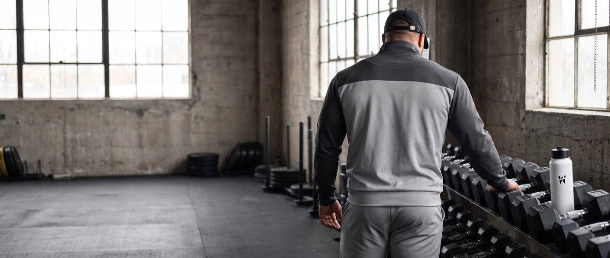 A man in the Grey Drift Set and WAYSHNER cap, seen from behind, by a dumbbell rack in a gym